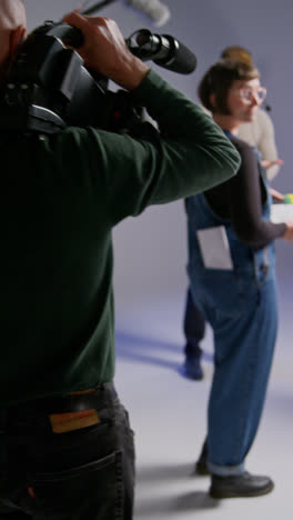 Vertical-Video-Of-Film-Cameraman-Shooting-Movie-Handheld-On-Busy-Film-Set-In-Studio-With-Sound-Recordist-And-Assistant